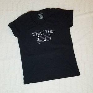 以下が含まれることがあります： 白い「WHAT THE」の文字と、トレブルクレフと音符が白でプリントされた、黒いVネックTシャツです。