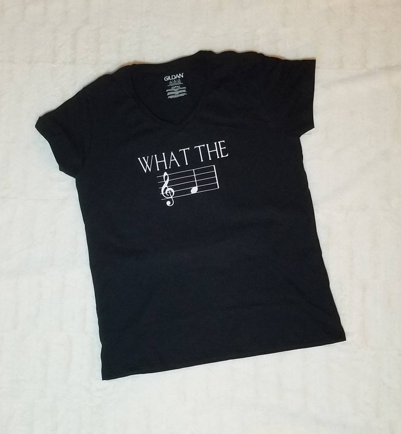 以下が含まれることがあります： 白い「WHAT THE」の文字と、トレブルクレフと音符が白でプリントされた、黒いVネックTシャツです。