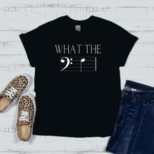 以下が含まれることがあります： 「WHAT THE」というテキストと五線譜に書かれた音符がプリントされた黒いTシャツ