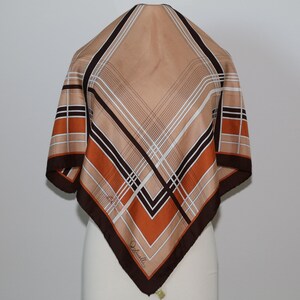 May include: A square scarf with a geometric pattern in shades of tan, brown, orange, and white. The scarf has a dark brown border and features a diagonal plaid design. The scarf is displayed on a mannequin.