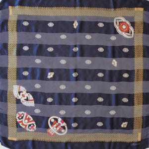 May include: A square silk scarf with a navy blue and gray striped pattern. The scarf features decorative embellishments in red, white, and gold, along with small white eye-shaped designs. The border has a gold and brown checkered pattern.