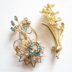 May include: Two vintage gold-tone brooches. One features a floral design with teal and clear rhinestones. The other is a bouquet of flowers. Both are set against a white background.