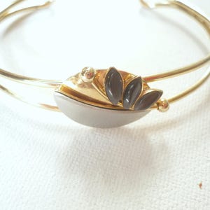 Könnte beinhalten: Ein Armreif aus goldfarbenem Metall mit einem zentralen Dekorelement. Dieses Mittelstück besteht aus einem glatten, geschwungenen silberfarbenen Abschnitt und einem goldfarbenen fächerförmigen Detail, das mit drei kleinen, dunklen, marquiseförmigen Steinen besetzt ist.