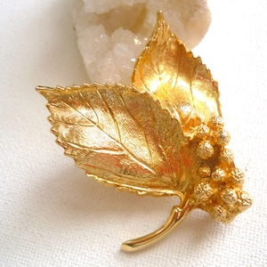 May include: A gold-toned leaf brooch with a cluster of small, round, textured details. The brooch is made of metal and has a realistic leaf design.
