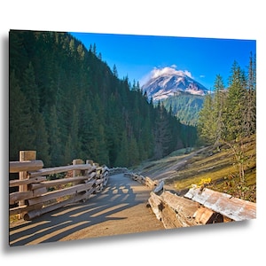 May include: A scenic landscape print featuring a mountain peak partially obscured by clouds. A wooden fence lines a paved path that winds through a valley surrounded by evergreen trees. The sky is a clear, bright blue.