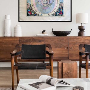 May include: A framed vintage illustration of a geocentric model of the solar system. The illustration is in a black frame and is hanging on a white wall above a wooden cabinet. The cabinet has two doors and is flanked by two chairs with black leather seats. A white marble coffee table sits in front of the cabinet with a book and a magazine on top.