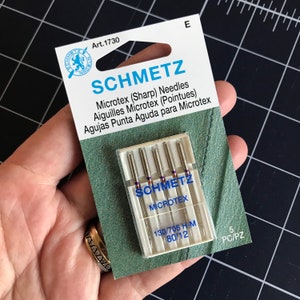 May include: A package of 5 Schmetz Microtex sewing machine needles in a clear plastic case. The needles are silver and the packaging is white with blue and purple text. The text on the package reads "Schmetz Microtex 130/705 H-M 80/12 5 PC/PZ".