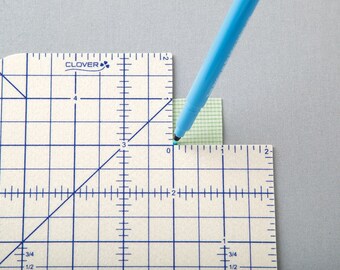 Può includere: Primo piano di una penna blu che segna un punto su un righello bianco a griglia con la scritta "CLOVER" stampata su di esso. Il righello è segnato in pollici e frazioni di pollice.