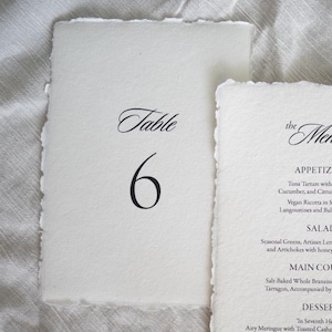 Peut inclure: Deux cartons en papier faits à la main. L'un lit « Table 6 » en écriture élégante. L'autre affiche un menu avec des entrées, une salade, un plat principal et des desserts. Le papier a un bord texturé et découpé.