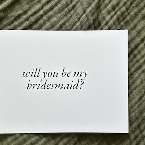 May include: A white card with the text "will you be my bridesmaid?" in a cursive font. The card is set against a textured, olive green fabric background. The card is rectangular and the text is centered.