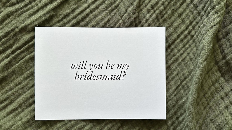 May include: A white card with the text "will you be my bridesmaid?" in a cursive font. The card is set against a textured, olive green fabric background. The card is rectangular and the text is centered.
