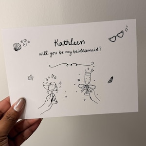 May include: A white rectangular card featuring black line art illustrations and the text "Kathleen will you be my bridesmaid?". The design includes drawings of two hands holding champagne glasses, a seashell, a small star, and sunglasses.