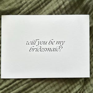 May include: A white card with the text "will you be my bridesmaid?" in elegant script. The card is set against a textured, olive green background, suggesting a wedding or event theme.