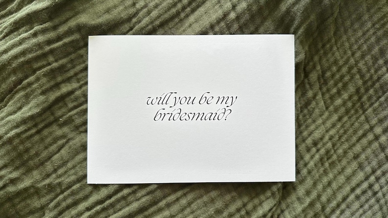 May include: A white card with the text "will you be my bridesmaid?" in elegant script. The card is set against a textured, olive green background, suggesting a wedding or event theme.