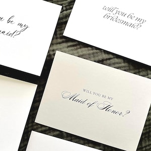 Puede incluir: Tarjetas blancas con caligrafía elegante que preguntan "Will you be my bridesmaid?" y "Will you be my Maid of Honor?" dispuestas sobre una superficie texturizada verde oscuro, creando un diseño simple y clásico.