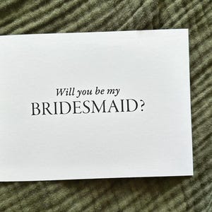 May include: A white card with the text "Will you be my BRIDESMAID?" in a simple, elegant font. The card is centered on a textured, olive green fabric background. Ideal for wedding proposals.