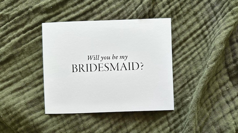 May include: A white card with the text "Will you be my BRIDESMAID?" in a simple, elegant font. The card is centered on a textured, olive green fabric background. Ideal for wedding proposals.