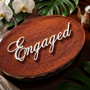 May include: An oval-shaped tiramisu dessert topped with a thick layer of cocoa powder and a white script cake topper that reads Engaged. The dessert is presented with silver cutlery, a silver teapot, and white orchid flowers nearby.