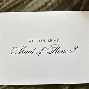 May include: A white card with the text "WILL YOU BE MY Maid of Honor?" in elegant black script. The card is rectangular and sits on a textured, olive-green surface, suggesting a wedding or bridal theme.