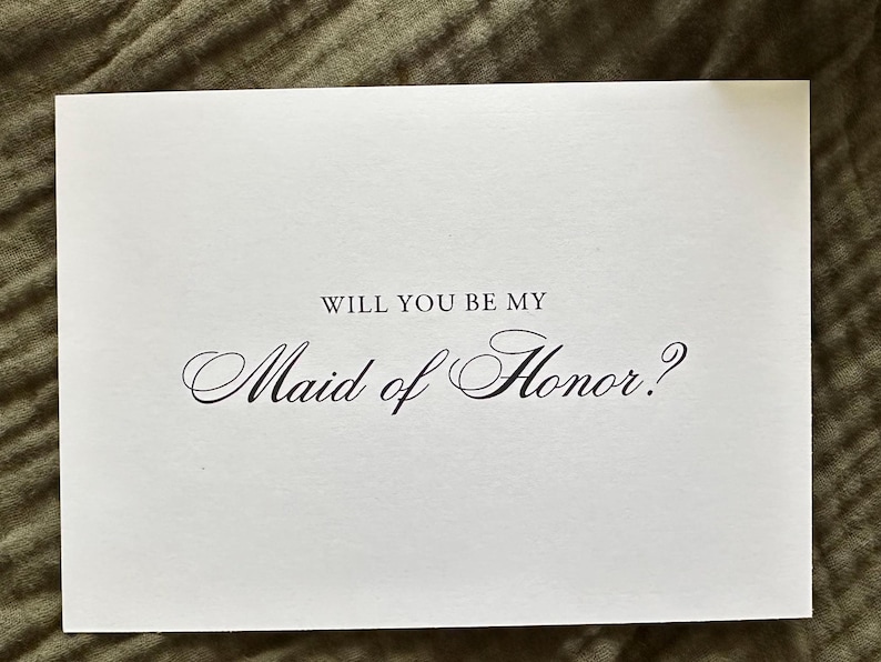 May include: A white card with the text "WILL YOU BE MY Maid of Honor?" in elegant black script. The card is rectangular and sits on a textured, olive-green surface, suggesting a wedding or bridal theme.
