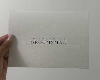 May include: A white card with the text "Devin, will you be my groomsman?" printed in black.