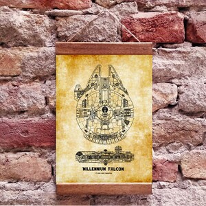 Może przedstawiać: Wizerunek w stylu vintage statku kosmicznego Millennium Falcon z Gwiezdnych Wojen. Druk na beżowym tle z brązową drewnianą ramą. Tekst "MILLENNIUM FALCON" jest nadrukowany na dole wydruku.