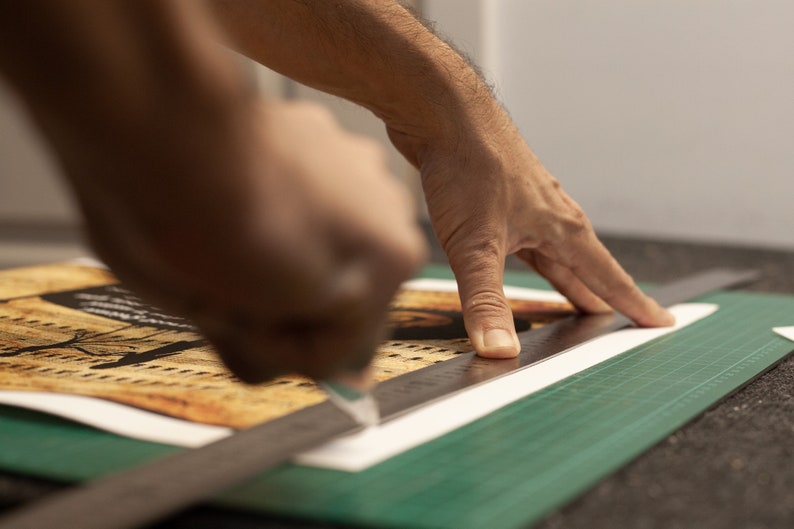 Puede incluir: Una mano utiliza una regla y un c&uacute;ter para cortar un trozo de papel con un dise&ntilde;o impreso en una estera de corte verde.