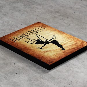 Könnte beinhalten: Rechteckiges Holzschild mit schwarzem Rand und einem verwitterten, sepiafarbenen Hintergrund. Das Schild zeigt die Silhouette einer Figur, die ein Getränk einschenkt, mit dem Wort "DRINKERBELL" in einer stilisierten Schriftart. Das Gesamtthema bezieht sich auf Trinken und Bars.