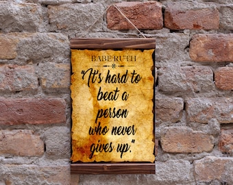 Op de afbeelding: Een houten bord met een vintage perkament achtergrond met een citaat van Babe Ruth: "Het is moeilijk om iemand te verslaan die nooit opgeeft."