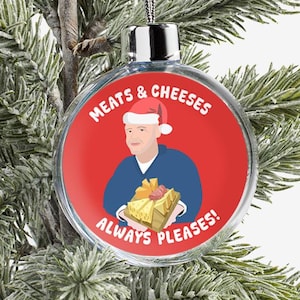 May include: A clear, round Christmas bauble with a red background and the words "MEATS & CHEESES ALWAYS PLEASES!" The bauble features an illustration of a person in a Santa hat holding a gift.