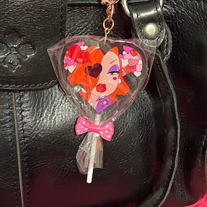 Puede incluir: Un bolso de cuero negro con un charm de piruleta en forma de corazón que presenta un personaje de dibujos animados con cabello rojo y un lazo rosa. La piruleta es transparente y tiene un palo blanco.