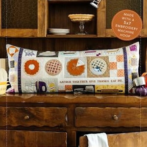 May include: A decorative bench pillow with pie-themed embroidery. The pillow features various pie slice designs in orange, pink, and blue, with the text "GATHER TOGETHER, GIVE THANKS, EAT PIE." The pillow is made with a 5x7 embroidery machine hoop.