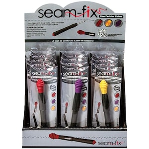 May include: A display of seam rippers with the text "seam-fix." The rippers have black handles and colored tips in red, purple, and yellow. The packaging includes the text "New Fashion Colors."