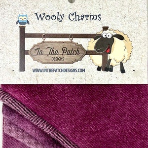May include: A folded fabric sample in shades of purple, with a patterned edge. The top portion features a decorative sign with the text "Wooly Charms" and "In The Patch Designs", along with a cartoon sheep and owl.