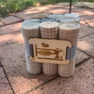 May include: A set of 9 wool felt squares in various shades of beige and gray, rolled up and stacked in a cube shape. The squares are held together with a wooden band that says "Cube" and "In The Patch Wool" with a website address.