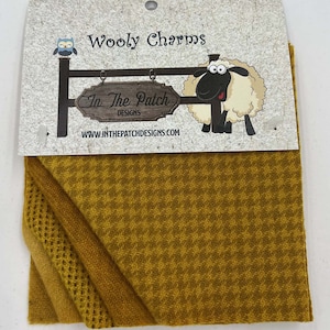 May include: A pack of three wool fabric squares in shades of yellow. The fabric is a herringbone pattern. The packaging features a cartoon sheep and an owl with the text "Wooly Charms" and "In The Patch Designs".