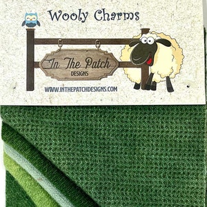 May include: Wooly Charms fabric squares in shades of green. The fabric is a woven wool blend with a subtle texture. The fabric squares are packaged with a label that says "Wooly Charms" and features a cartoon sheep and owl.