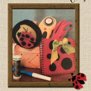 May include: A red felt pincushion shaped like a ladybug with black spots and a yellow ribbon. The pincushion is in a basket with other sewing supplies, including a spool of black thread, a pair of scissors, and a rotary cutter. The image includes the text "Lady Bug Tie On Pincushion & Thread Catcher".