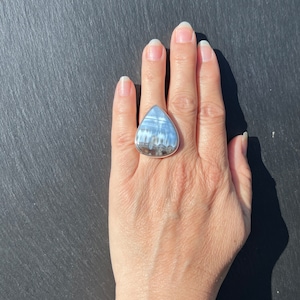 May include: A silver ring with a large, triangular, white and gray gemstone. The ring is on a person's finger.