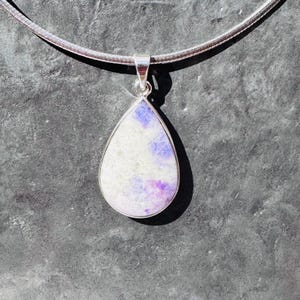 May include: A teardrop-shaped pendant with a white and purple stone set in a silver frame. The pendant hangs from a silver chain, creating a simple yet elegant necklace. The stone has a speckled pattern.