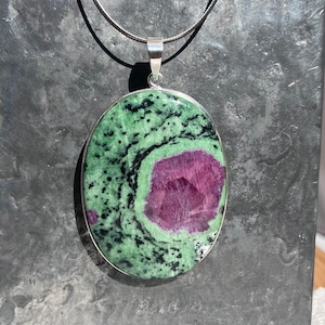 Può includere: Una collana con catena d'argento con un ciondolo ovale con una pietra verde e nera con un centro rosa.