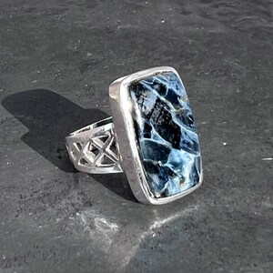 Op de afbeelding: Een zilveren ring met een blauwe en witte tijgeroog-steen gezet in een rechthoekige bezel. De band is een delicaat, opengewerkt ontwerp.