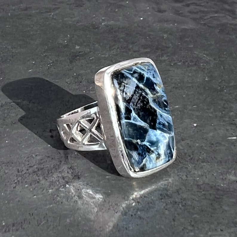 Op de afbeelding: Een zilveren ring met een blauwe en witte tijgeroog-steen gezet in een rechthoekige bezel. De band is een delicaat, opengewerkt ontwerp.