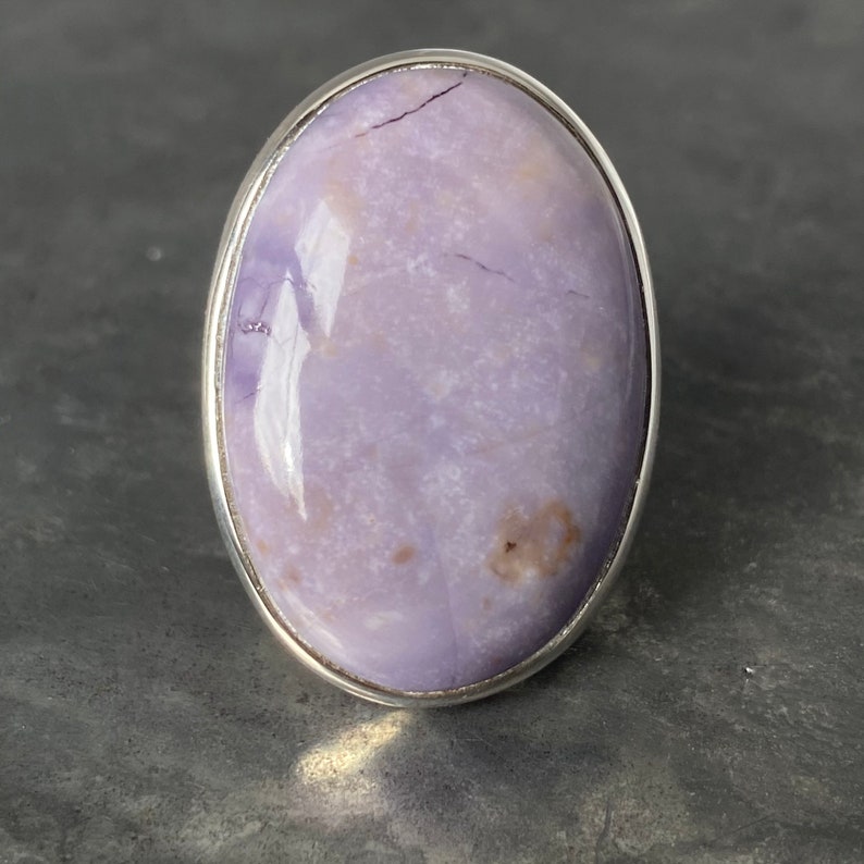 May include: A silver ring with a large oval-shaped purple gemstone. The gemstone has a marbled pattern and a shiny finish.