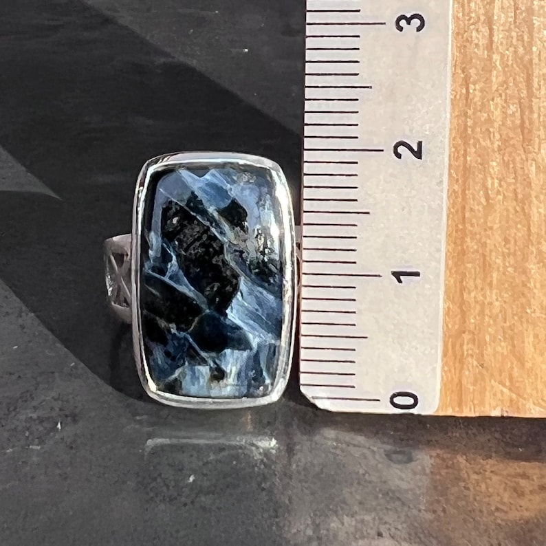 Op de afbeelding: Een zilveren ring met een rechthoekige blauwe en zwarte tijgeroog-edelsteen. De ring ligt naast een liniaal, wat aangeeft dat de steen ongeveer 2,5 cm lang is.