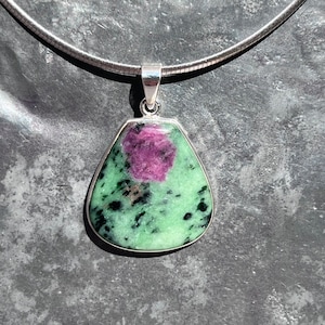 May include: A silver pendant necklace featuring a green and pink gemstone with black flecks. The gemstone is set in a silver bezel and hangs from a silver chain.