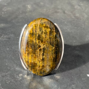 May include: A silver ring with a large oval-shaped tiger's eye gemstone. The stone has a brown and gold color with a striated pattern.