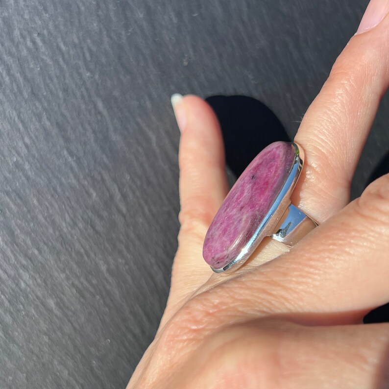Peut inclure: Une bague ovale avec une pierre pr&eacute;cieuse rose vif sertie dans un anneau en m&eacute;tal argent&eacute;. La pierre a un aspect textur&eacute; avec des inclusions roses plus fonc&eacute;es. La bague est port&eacute;e sur un doigt, sur fond sombre.