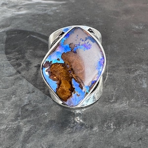 May include: A wide silver ring featuring a large, oval-shaped opal. The opal displays a vibrant mix of colors, including blues, browns, and hints of purple. The ring's band is thick and has a textured appearance, adding to its unique design.