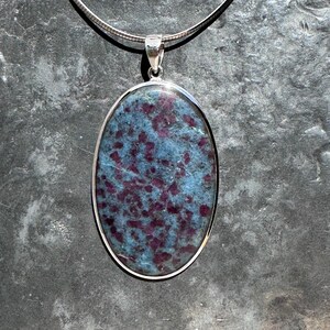 Puede incluir: Un colgante ovalado de plata con una piedra preciosa azul y morada. La piedra preciosa tiene un patrón moteado.